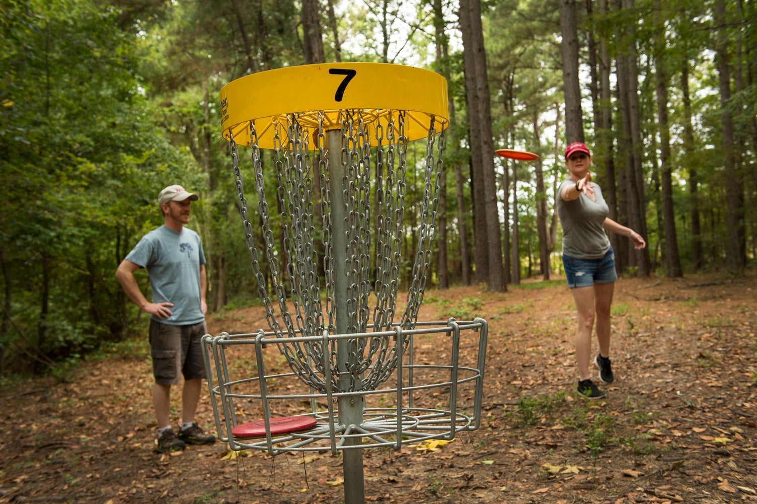 Woman standing behind disc golf basket while tossing a disc into it, man standing beside her watching 